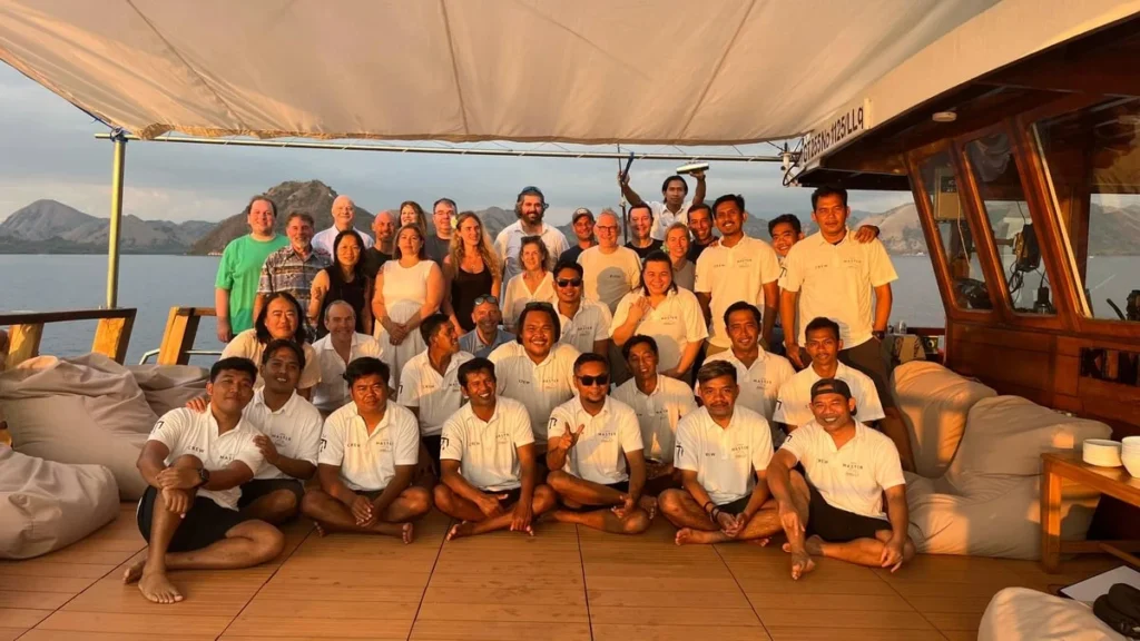 Group photo of guests and crew aboard Indo Master, smiling together on deck during a sunset in Indonesia, showcasing the camaraderie and family-like atmosphere of the liveaboard experience
