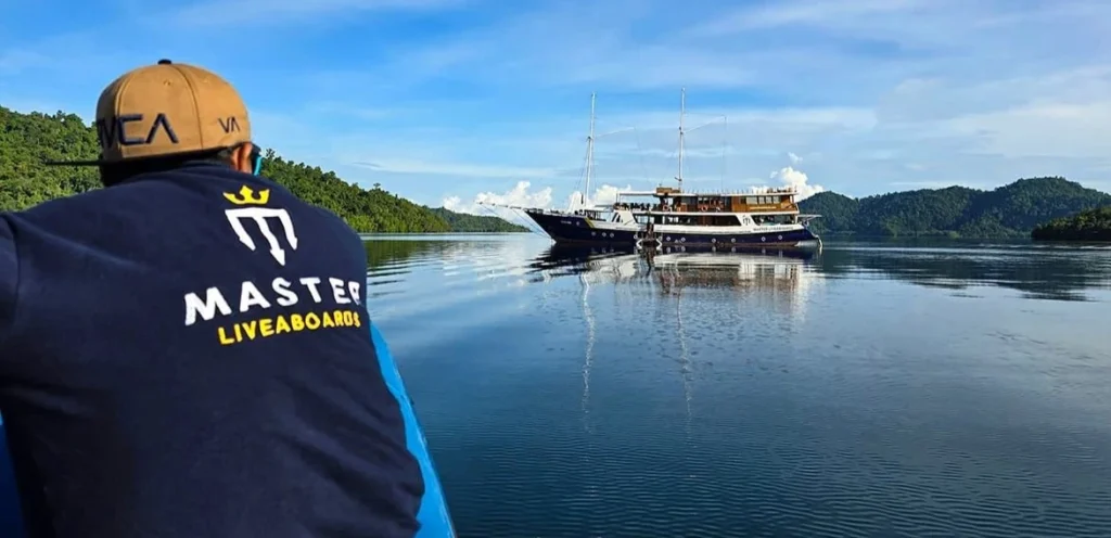 “Crew member wearing a Master Liveaboards uniform on board Indo Master, with the scenic islands of Raja Ampat in the background.