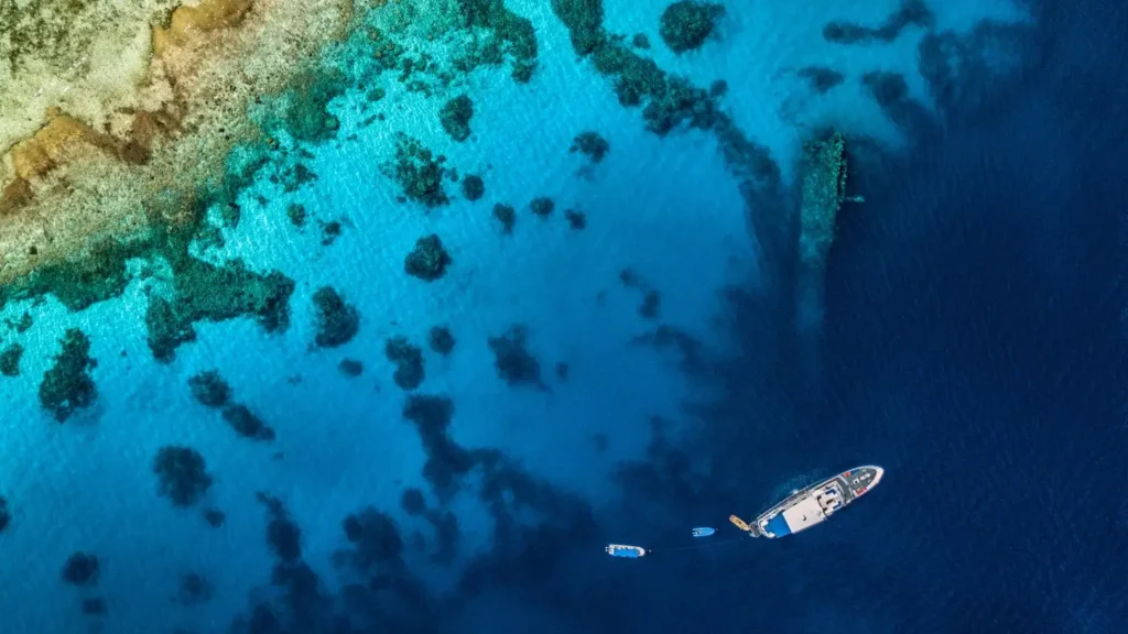 Wide-angle shot of a liveaboard vessel anchored in clear turquoise water above a large shipwreck, illustrating the isolation and the need for self-contained sustainability.